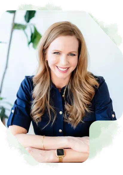Heather Siegel specializes in web design and SEO in Highlands Ranch CO. She has red hair, blue eyes, is wearing a navy blue dress and is sitting at a desk with plants in the background